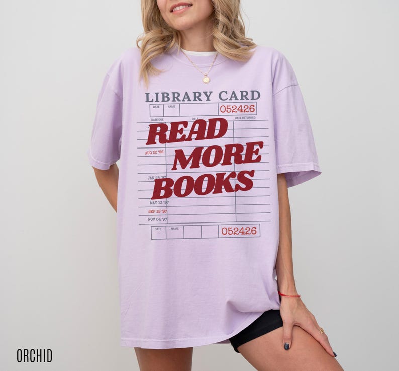 Woman wearing an oversized orchid Comfort Colors t shirt with a retro library card design and bold red Read More Books text, styled with black biker shorts and gold jewelry for a cozy book lover aesthetic