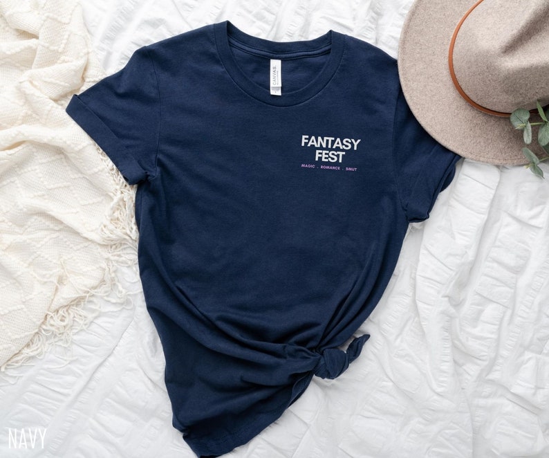 May include: A navy blue t-shirt with the text "Fantasy Fest" in white with the words "Magic, Romance, Smut" in smaller purple text below.