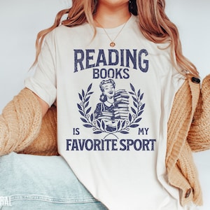 May include: Cream-colored t-shirt with the text "READING BOOKS IS MY FAVORITE SPORT" in navy blue. The design includes a vintage illustration of a woman holding books, surrounded by a laurel wreath. The word "NATURAL" is printed at the bottom left.