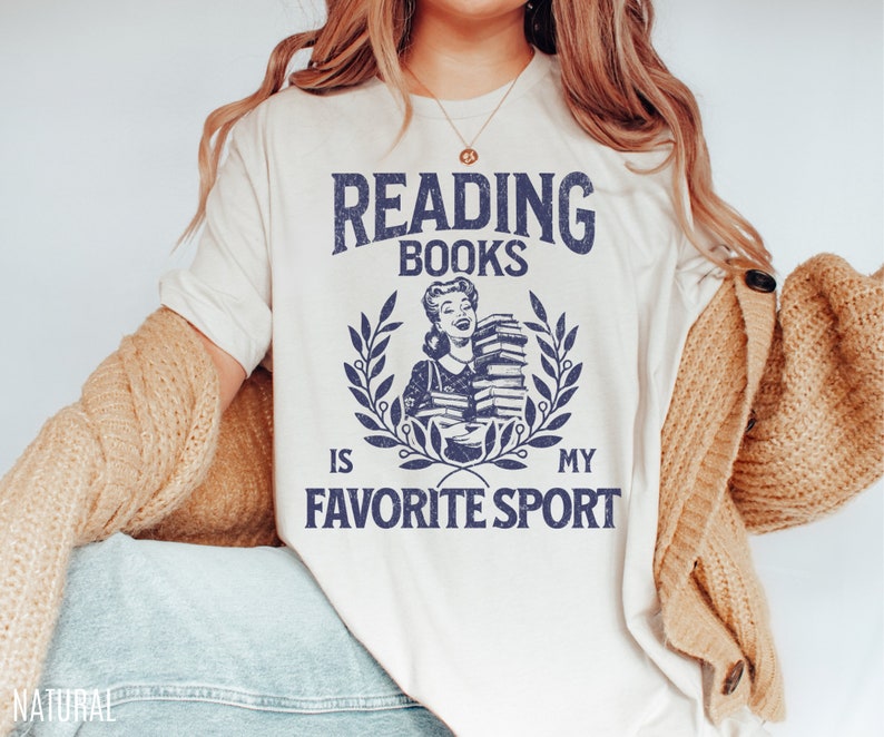 May include: Cream-colored t-shirt with the text "READING BOOKS IS MY FAVORITE SPORT" in navy blue. The design includes a vintage illustration of a woman holding books, surrounded by a laurel wreath. The word "NATURAL" is printed at the bottom left.