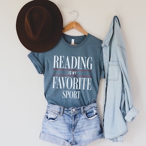 Może przedstawiać: Niebieska koszulka z napisem "Reading is my favorite sport" białymi literami z czerwonymi liniami. Koszulka wisi na drewnianym wieszaku z brązowym kapeluszem i jasnoniebieską koszulą dżinsową.