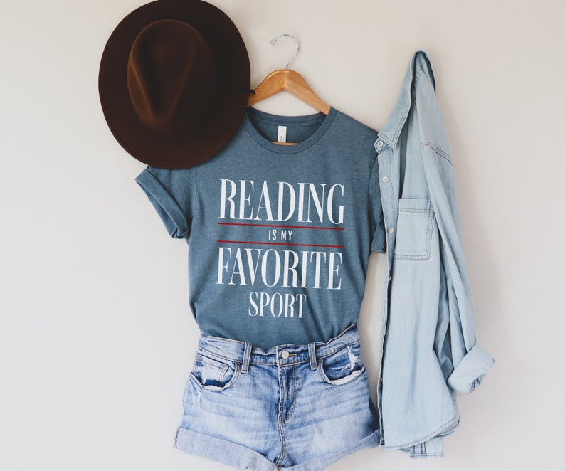 Może przedstawiać: Niebieska koszulka z napisem "Reading is my favorite sport" białymi literami z czerwonymi liniami. Koszulka wisi na drewnianym wieszaku z brązowym kapeluszem i jasnoniebieską koszulą dżinsową.