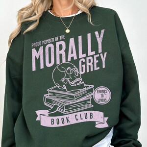 Puede incluir: Una sudadera verde oscuro con un estampado gr&aacute;fico rosa que dice "Proud Member of the Morally Grey Book Club". El gr&aacute;fico tambi&eacute;n incluye una calavera, libros y el texto "Enemies to Lovers".