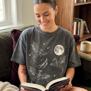 May include: Dark grey t-shirt with a fantasy design of dragons, a full moon, and shooting stars. The shirt has a vintage wash appearance. The person is reading a book titled "Stargazer's Apprentice".
