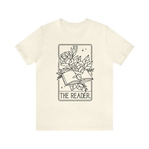 May include: Cream-colored t-shirt featuring a black graphic of an open book with roses and leaves, framed by a rectangle with the Roman numeral XII and stars. The text "THE READER." is below the book.