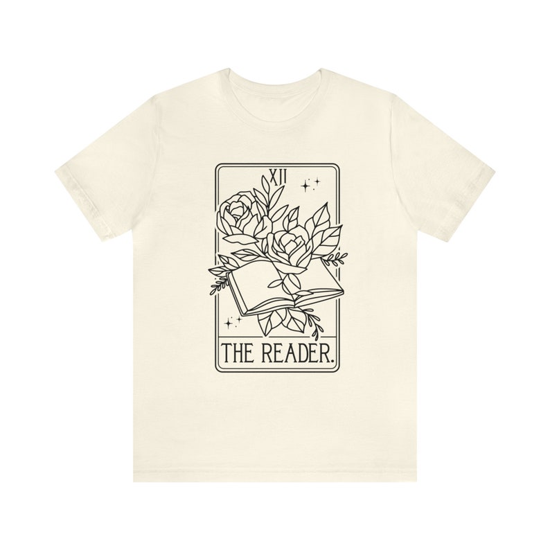 May include: Cream-colored t-shirt featuring a black graphic of an open book with roses and leaves, framed by a rectangle with the Roman numeral XII and stars. The text "THE READER." is below the book.