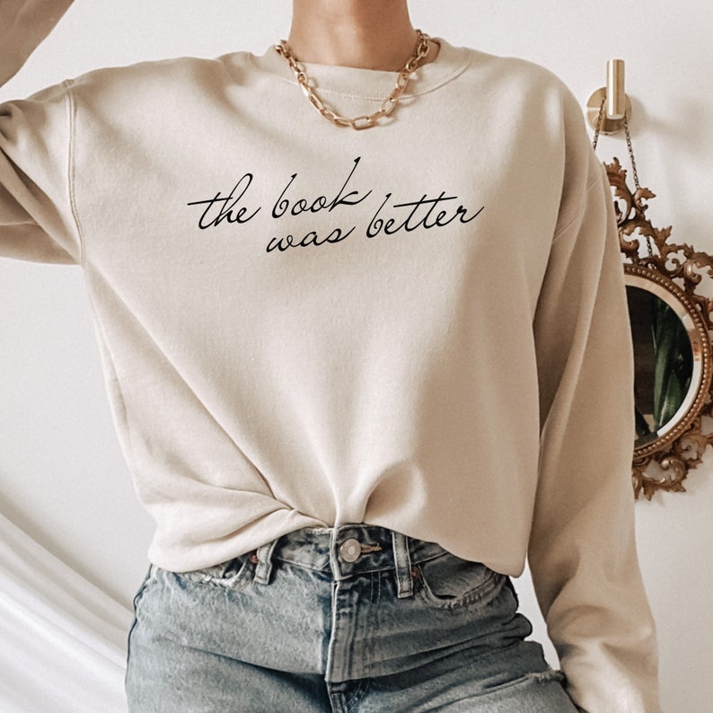 Puede incluir: Una sudadera de cuello redondo en color beige con el texto en cursiva negra "the book was better" impreso en el pecho. La prenda tiene mangas largas y cuello acanalado, combinada con pantalones vaqueros azules y un collar de cadena dorada.
