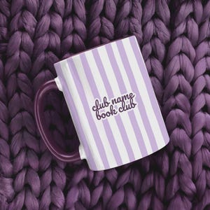 Puede incluir: Una taza de cerámica blanca con rayas verticales lavanda y un asa morada oscura. La taza presenta el texto "club name book club" en cursiva. La taza está colocada sobre un fondo tejido morado oscuro texturizado.
