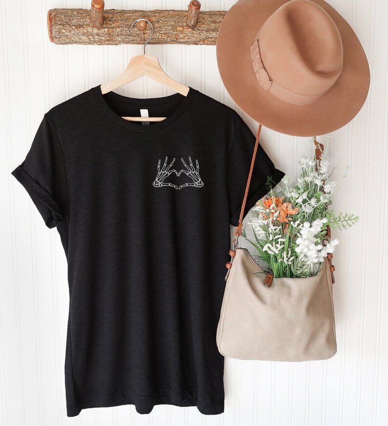 May include: Black t-shirt with a white skeleton hand heart graphic. The shirt is hanging on a wooden hanger. A tan hat and a beige purse with flowers are also visible.