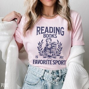 May include: Light pink t-shirt with the text "READING BOOKS IS MY FAVORITE SPORT" in navy blue. The graphic features a woman holding books within a laurel wreath. The shirt is a crew neck and has short sleeves.