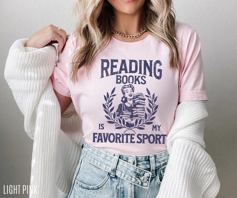 May include: Light pink t-shirt with the text "READING BOOKS IS MY FAVORITE SPORT" in navy blue. The graphic features a woman holding books within a laurel wreath. The shirt is a crew neck and has short sleeves.