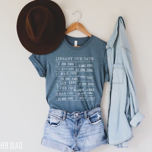 May include: A blue t-shirt with a white graphic that says "Library Due Date" and lists dates from 1980 to 1982. The shirt is hanging on a wooden hanger.