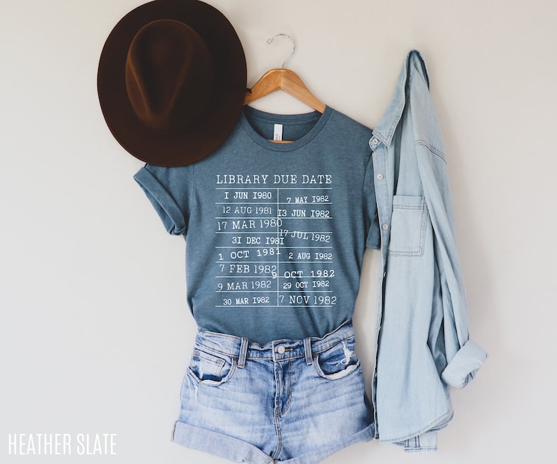 May include: A blue t-shirt with a white graphic that says "Library Due Date" and lists dates from 1980 to 1982. The shirt is hanging on a wooden hanger.