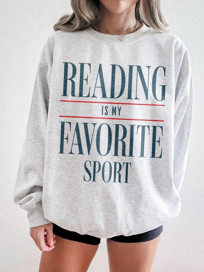 Woman wearing an ash sweatshirt featuring a vintage style graphic that says "reading is my favorite sport"