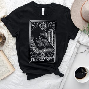 May include: Black t-shirt featuring a white graphic of an open book, stacks of books, a sun, and stars, with the text "THE READER." The design is framed within a rectangular border. A hat and a cup of coffee are in the background.