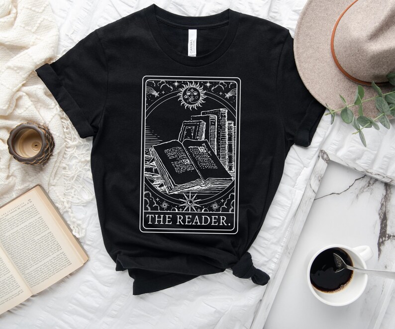 May include: Black t-shirt featuring a white graphic of an open book, stacks of books, a sun, and stars, with the text "THE READER." The design is framed within a rectangular border. A hat and a cup of coffee are in the background.