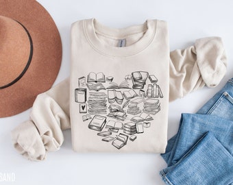 May include: A sand-colored sweatshirt featuring a heart-shaped design composed of various sketched books. The design includes open and closed books, and stacks of books. A brown hat and blue jeans are also in the image.