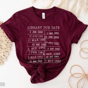 May include: A maroon t-shirt with white text that reads "LIBRARY DUE DATE" and lists dates from 1980 to 1982.