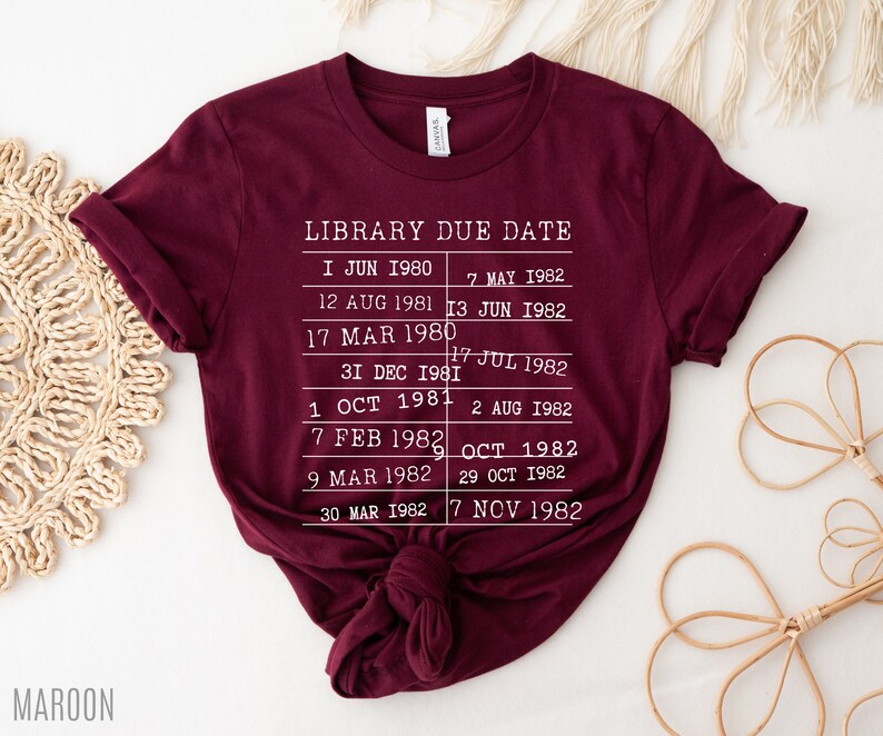 May include: A maroon t-shirt with white text that reads "LIBRARY DUE DATE" and lists dates from 1980 to 1982.