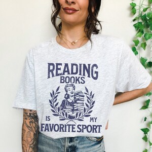 May include: Light gray t-shirt with the text "READING BOOKS IS MY FAVORITE SPORT" in navy blue. The graphic features a woman holding books within a laurel wreath. The shirt is a casual, comfortable style.