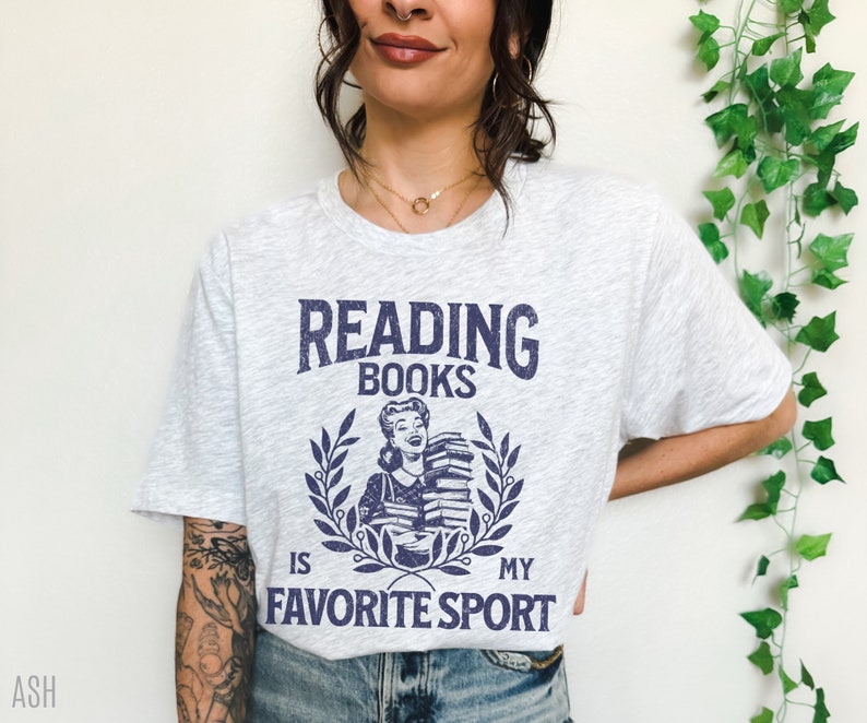 May include: Light gray t-shirt with the text "READING BOOKS IS MY FAVORITE SPORT" in navy blue. The graphic features a woman holding books within a laurel wreath. The shirt is a casual, comfortable style.