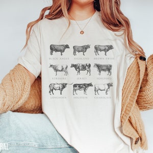 Puede incluir: Camiseta color crema con ilustraciones detalladas de nueve razas de vacas, incluyendo Black Angus y Jersey. Cada vaca est&aacute; etiquetada con el nombre de su raza en una fuente cl&aacute;sica. La palabra "NATURAL" est&aacute; impresa en la parte inferior izquierda.