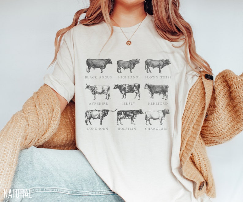 Puede incluir: Camiseta color crema con ilustraciones detalladas de nueve razas de vacas, incluyendo Black Angus y Jersey. Cada vaca est&aacute; etiquetada con el nombre de su raza en una fuente cl&aacute;sica. La palabra "NATURAL" est&aacute; impresa en la parte inferior izquierda.