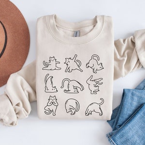 May include: A cream-coloured sweatshirt with a black line drawing of cats performing yoga. The design shows cats in poses such as downward dog and lotus. The sweatshirt is folded on a white surface with a brown hat and blue jeans.