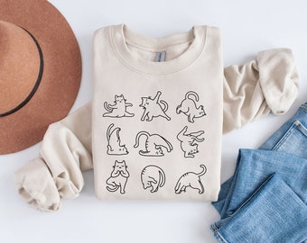 May include: A beige sweatshirt featuring a black line drawing of cats in various yoga poses. The design includes cats in poses like downward dog and lotus position. The sweatshirt is folded on a white surface with a brown hat and blue jeans.