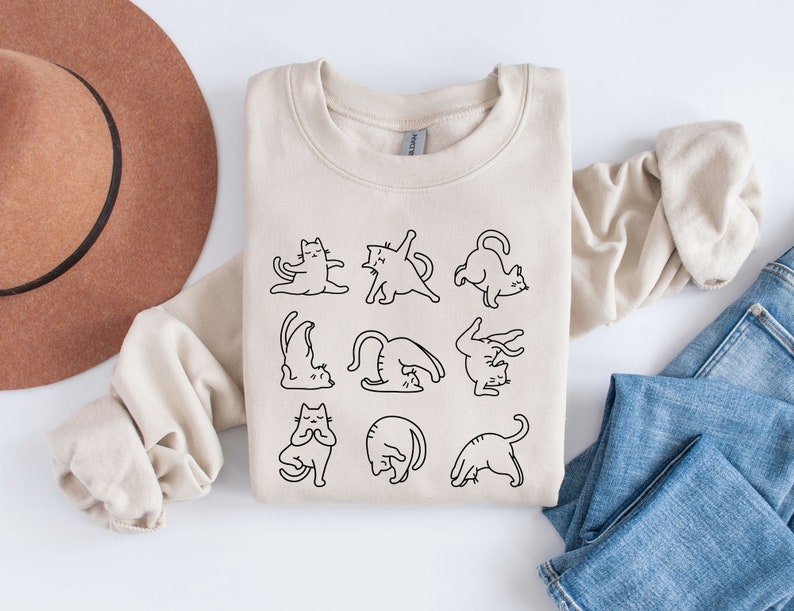 May include: A cream-coloured sweatshirt with a black line drawing of cats performing yoga. The design shows cats in poses such as downward dog and lotus. The sweatshirt is folded on a white surface with a brown hat and blue jeans.