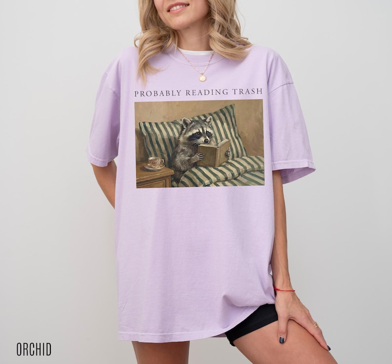 Woman wearing oversized orchid purple t shirt with probably reading trash text and raccoon reading book design, styled casually
