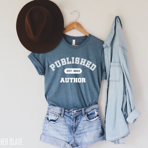 May include: A teal blue t-shirt with white text that reads "PUBLISHED EST. 2022 AUTHOR". The shirt is hanging on a wooden hanger and is styled with denim shorts and a light blue denim jacket.