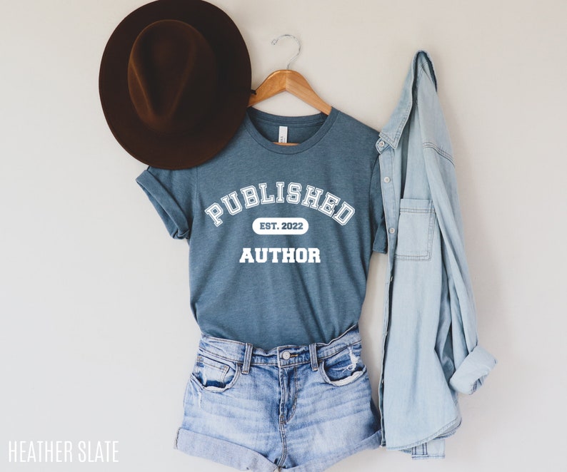 May include: A teal blue t-shirt with white text that reads "PUBLISHED EST. 2022 AUTHOR". The shirt is hanging on a wooden hanger and is styled with denim shorts and a light blue denim jacket.