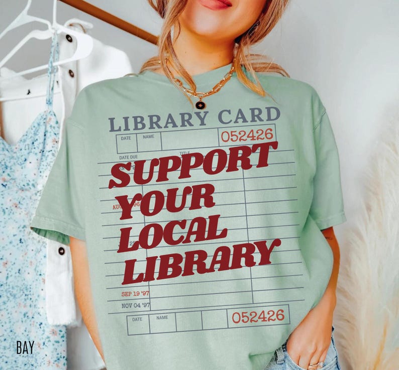 Woman wearing oversized bay green t shirt with vintage library card graphic and support your local library text styled indoors