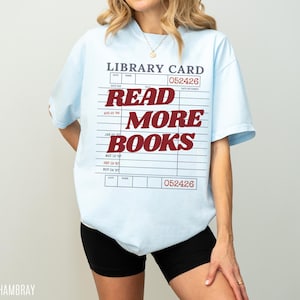 Woman wearing an oversized chambray blue Comfort Colors t shirt featuring a vintage library card design with bold red Read More Books lettering, paired with black biker shorts and gold jewelry for a casual reader inspired outfit and literary aesthetic look