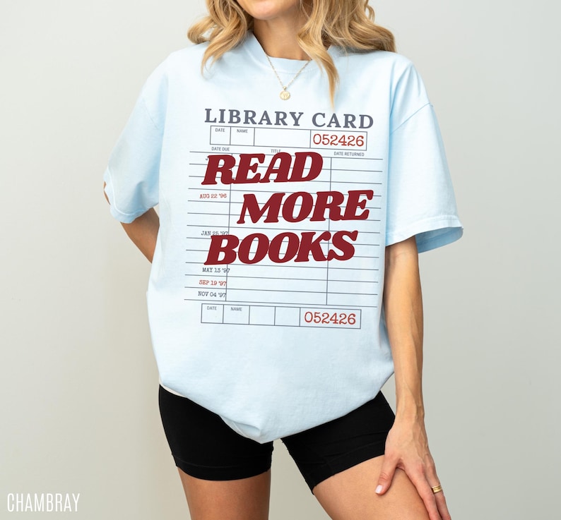 Woman wearing an oversized chambray blue Comfort Colors t shirt featuring a vintage library card design with bold red Read More Books lettering, paired with black biker shorts and gold jewelry for a casual reader inspired outfit and literary aesthetic look
