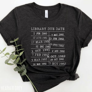 May include: Dark heather grey t-shirt with a white graphic of a library due date list. The list includes dates from June 1980 to November 1982.
