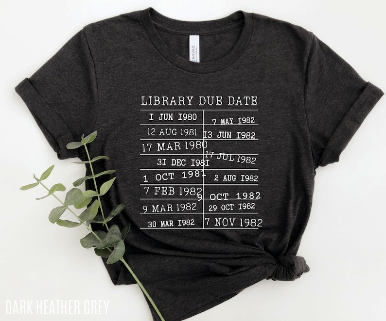 May include: Dark heather grey t-shirt with a white graphic of a library due date list. The list includes dates from June 1980 to November 1982.
