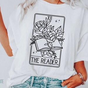 May include: White t-shirt featuring a black and white graphic of a tarot card. The card depicts an open book with roses and the words "THE READER." The shirt also has the word "WHITE" printed on the bottom left.
