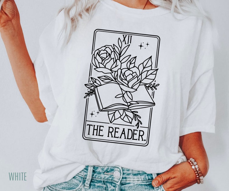 May include: White t-shirt featuring a black and white graphic of a tarot card. The card depicts an open book with roses and the words "THE READER." The shirt also has the word "WHITE" printed on the bottom left.