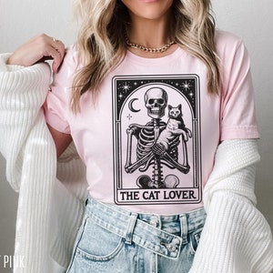May include: Light pink t-shirt featuring a black and white graphic of a skeleton holding a cat, with the text "THE CAT LOVER" below. The design is framed by a decorative border with stars and a crescent moon. The shirt is made of a soft material.