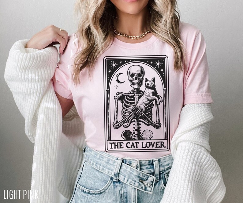 May include: Light pink t-shirt featuring a black and white graphic of a skeleton holding a cat, with the text "THE CAT LOVER" below. The design is framed by a decorative border with stars and a crescent moon. The shirt is made of a soft material.