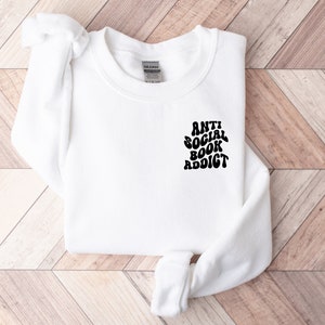 May include: White sweatshirt with the text "ANTI SOCIAL BOOK ADDICT" in black, stylized font. The sweatshirt is laid flat on a light brown and white wooden surface. The sweatshirt has long sleeves and a crew neck.
