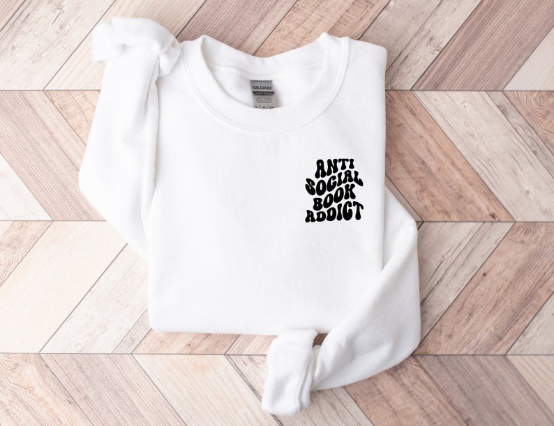 May include: White sweatshirt with the text "ANTI SOCIAL BOOK ADDICT" in black, stylized font. The sweatshirt is laid flat on a light brown and white wooden surface. The sweatshirt has long sleeves and a crew neck.