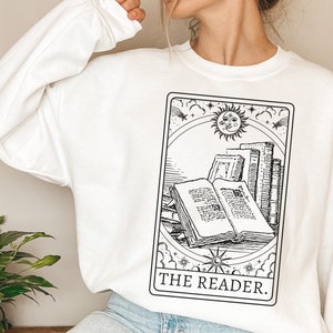 Puede incluir: Sudadera blanca con una ilustraci&oacute;n en blanco y negro de una carta de tarot que representa una pila de libros con el texto "THE READER."