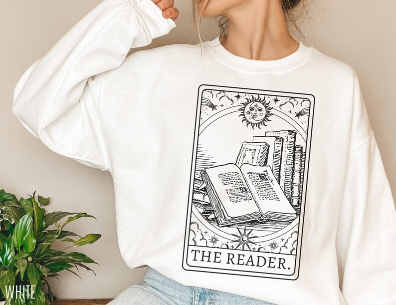 Puede incluir: Sudadera blanca con una ilustraci&oacute;n en blanco y negro de una carta de tarot que representa una pila de libros con el texto "THE READER."