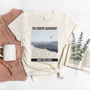 K&ouml;nnte beinhalten: Ein naturfarbenes T-Shirt mit einem grafischen Design, das eine Bergkette und den Text "The Riders Quadrant, Basgiath, War College" zeigt.