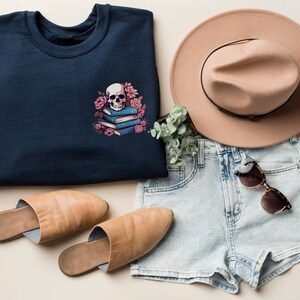 May include: A navy blue sweatshirt with a skull and floral design, alongside a tan hat, denim shorts, sunglasses, and tan slippers. The sweatshirt features a white skull on blue books with pink flowers. A casual, stylish outfit.