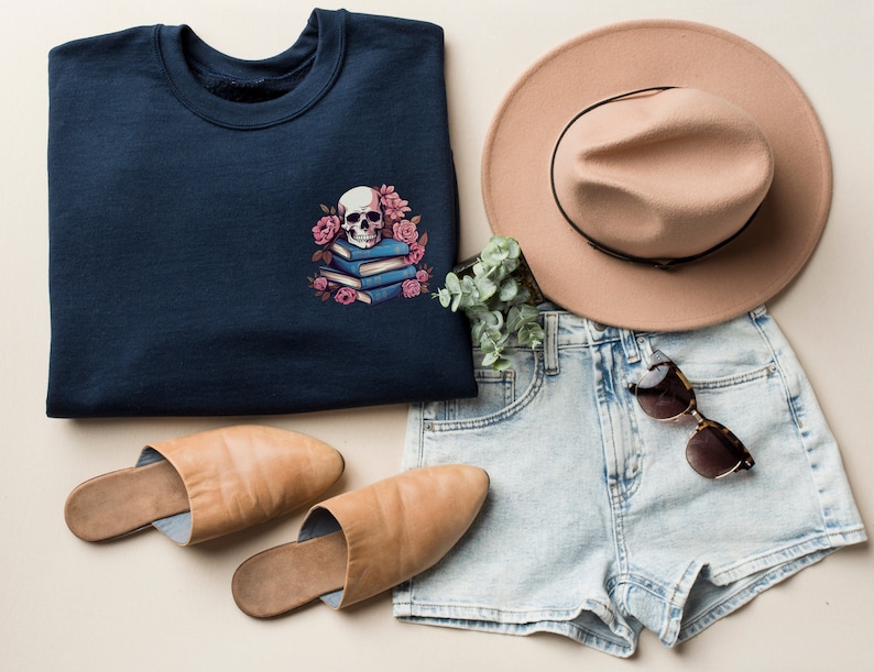 May include: A navy blue sweatshirt with a skull and floral design, alongside a tan hat, denim shorts, sunglasses, and tan slippers. The sweatshirt features a white skull on blue books with pink flowers. A casual, stylish outfit.