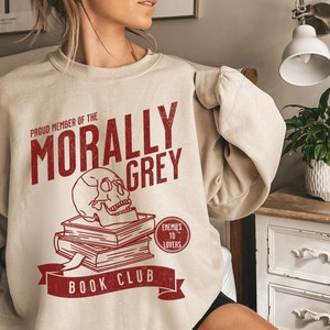 Puede incluir: Una sudadera beige con un dise&ntilde;o gr&aacute;fico rojo que dice "Proud Member of the Morally Grey Book Club". El gr&aacute;fico tambi&eacute;n incluye una calavera encima de una pila de libros y un c&iacute;rculo rojo que dice "Enemies to Lovers".